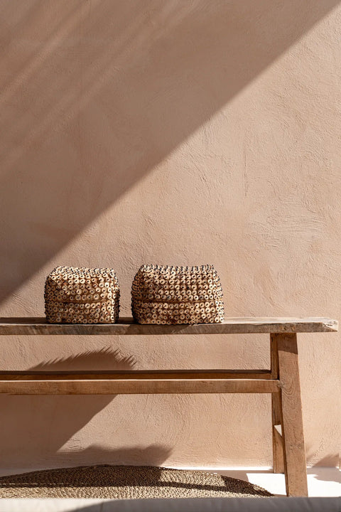 Two woven baskets, crafted by Balinese artisans, sit on a simple wooden bench against a textured, light beige wall. Sunlight casts a diagonal shadow across the scene, creating a calm and minimalist atmosphere.