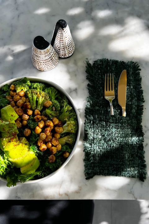 A bowl of salad with kale, broccoli, avocado slices, and chickpeas sits on a marble surface. Nearby, dining table accessories include a fork and knife on a textured green napkin, complemented by elegant salt and pepper sets in the background.