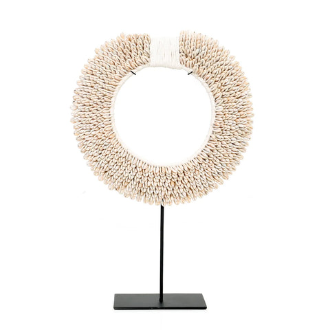 A decorative circular necklace made of numerous small, light-colored shells intricately arranged in rows. This statement piece is mounted on a black metal stand, bringing a coastal feel to any interior room with its elegant simplicity against a plain white background.