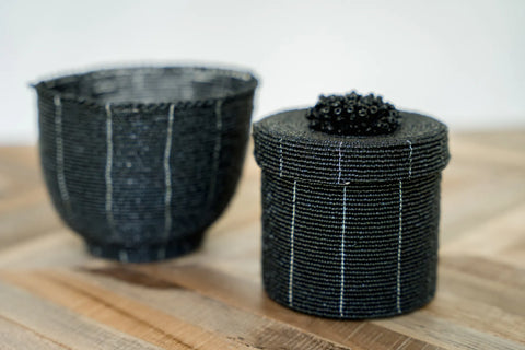 Two handcrafted black beaded containers grace a wooden surface, embodying boho chic decorative style. The left one is bowl-shaped, while the cylindrical piece on the right features a lid adorned with intricate bead details. Both handmade baskets showcase vertical white stripes, perfect for enhancing any home interior.