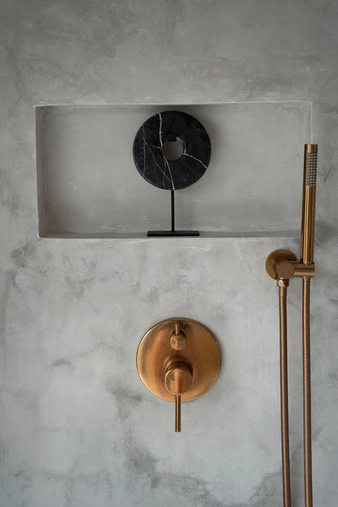 In a stylish home interior, a minimalist bathroom wall showcases a bronze shower handle against concrete. Above, a marble disc is elegantly displayed on a stand in the recessed shelf, enhancing the decorative display.