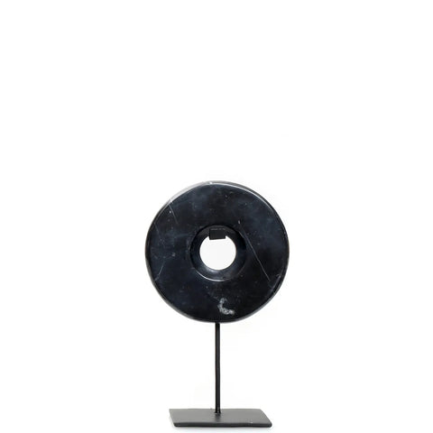 A black circular stone sculpture, reminiscent of a Marble Disc, with a center hole is mounted on a slender metal stand with a square base. Set against a plain white background, it serves as an elegant decorative display for any home interior.