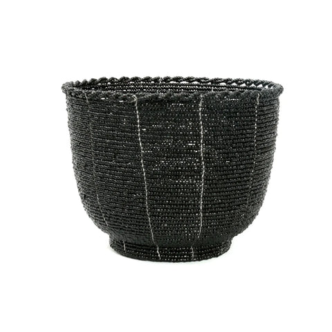 A round, intricately woven black basket with a textured surface and subtle vertical white lines adds a boho chic decorative touch to any home interior. Featuring a firm base and scalloped rim, this handmade beaded basket showcases detailed craftsmanship.