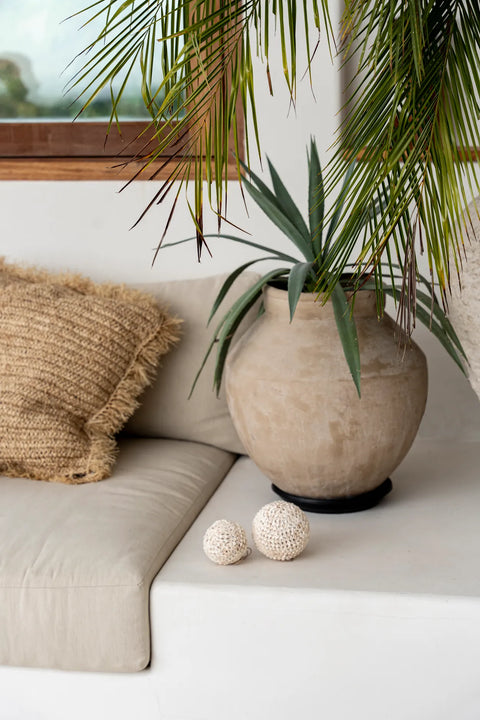 A cozy nook featuring a neutral-toned cushion with a textured pillow. Beside it, a large ceramic pot with green foliage complements the hand-made woven balls on the white surface, creating a minimalist and serene bohemian interior.