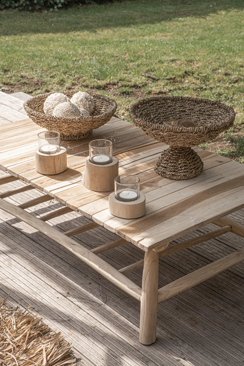 A wooden outdoor table on a deck showcases decorative wicker bowls with textured spheres, including rose-shaped shell balls crafted by Balinese craftsmen. Three glass candle holders complete the scene, while the background features a grassy lawn, creating a serene and natural setting.