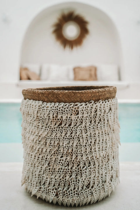 A hand-made woven basket with intricate white detailing stands in the foreground. In the background, a serene poolside area features cushioned seating and a decorative wall piece adorned with Balinese shells. The setting conveys a relaxed and stylish ambiance.