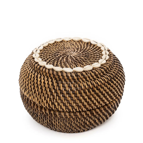 A round, rattan storage basket with a lid, made of natural brown fibers. The lid features a circular pattern of white cowrie shells around the edge, giving it a textured, rustic appearance.