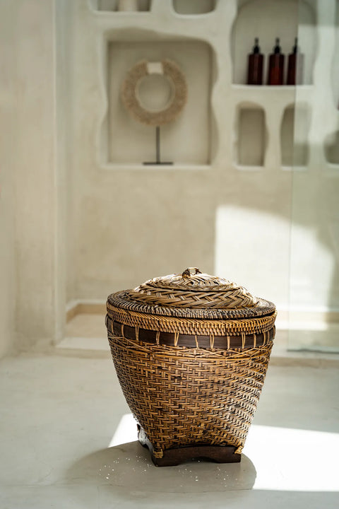 A woven wicker rattan basket with a lid rests on a light-colored floor. Sunlight streams in, casting a shadow on the ground. The background features a textured wall with niche shelves holding decorative home decor items, offering stylish storage solutions.