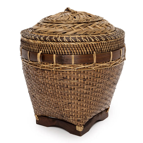 This tall, round woven basket, crafted from rattan, boasts a tightly fitted lid and intricate patterns in shades of brown. Resting on a small wooden base, it serves as both a charming piece of home decor and a practical storage solution with its traditional rustic style.