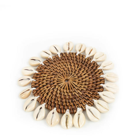 A round, woven wicker placemat offers a tropical touch with a border of cowrie seashells. The shells are evenly spaced around the edge, alternating in orientation on a plain white background.