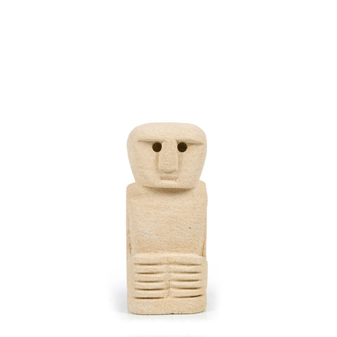 A beige sandstone sculpture of a simplified human figure with a rectangular head and small, circular eyes. The hand-carved piece showcases minimal facial features and detailed hands resting on its lap against a plain white background.