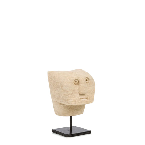 A hand-carved sandstone sculpture from the Sumba Stone collection features a stylized human face with large round eyes and a simple mouth, elegantly mounted on a black stand against a plain white background.