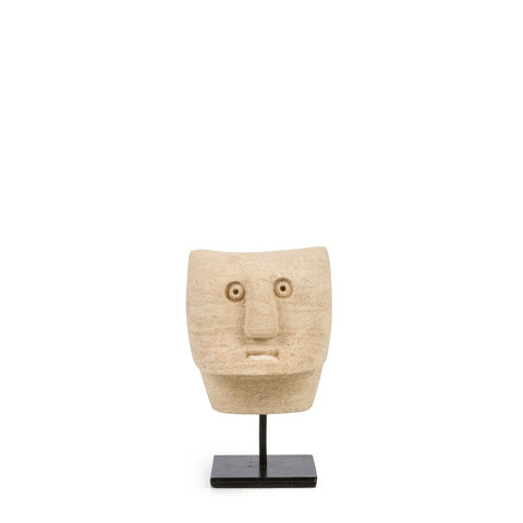 A hand-carved sandstone sculpture of a stylized face with wide eyes and a prominent nose, part of the Sumba Stone collection, is mounted on a black stand against a plain white background.