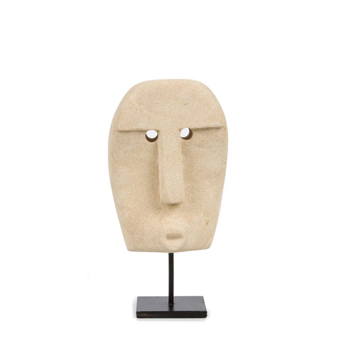 A beige, minimalist stone sculpture of a face with a long nose and small circular eyes, hand carved from sandstone, is mounted on a black stand. This piece draws inspiration from traditional Sumba statues and is set against a plain white background.