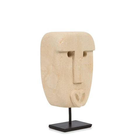 A hand-carved sandstone sculpture of a stylized human face, featuring minimalist elements like small eyes, a prominent nose, and a heart-shaped mouth. This piece reflects the craftsmanship of traditional Sumba artisans and is elegantly mounted on a black stand.