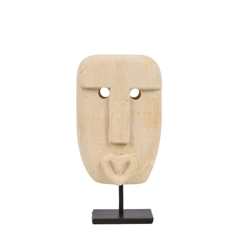 A beige stone mask, hand-carved from sandstone by traditional Sumba artisans, features a rectangular shape with circular eye holes, a long nose, and a stylized mouth. Mounted on a black metal stand, it stands against a plain white background.