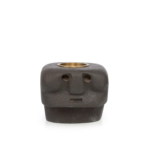 A small, square, black stone object resembling a stylized human face with angular features. It features a round brass insert on top, possibly for holding tealight holders or incense—a modern twist inspired by Sumba Stone statues.