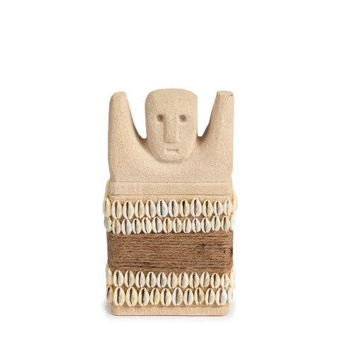 A beige clay sculpture with a simple human face and raised arms, reminiscent of hand-carved statues, is decorated with rows of cowrie shells and brown twine around its body. The piece stands against a plain white background.