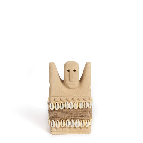 A small beige sculpture from the Sumba Stone collection features a simplistic human face and raised arms, adorned with rows of cowrie shells wrapped in twine. This unique statue sits against a plain white background, highlighting its hand-carved details.