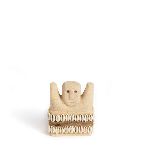 A small, beige Sumba Stone sculpture with a minimalistic human face and raised arms, adorned with rows of cowrie shells around its base, is meticulously hand-carved from sandstone. This exquisite piece is set against a plain white background, highlighting its delicate features.