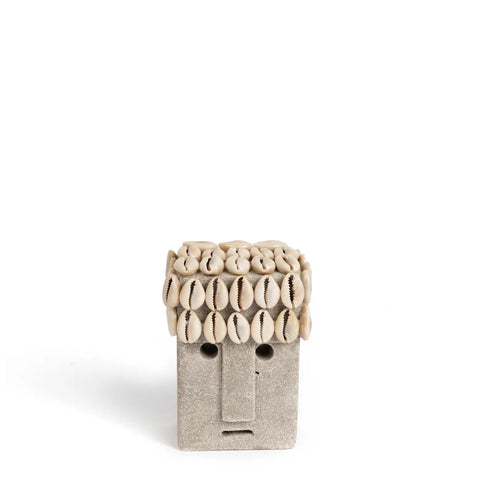 A small, minimalist Sumba Stone sculpture features a rectangular face with a flat expression. Hand carved with precision, its top is adorned with beige, shell-like shapes resembling hair. The background is a pristine white, highlighting the beauty of this unique piece.