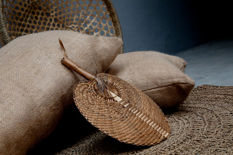 A woven hand fan rests on burlap-covered pillows atop a Seagrass Carpet. The background features parts of a wicker chair against a muted, dark wall in natural colors, creating a cozy, rustic ambiance.