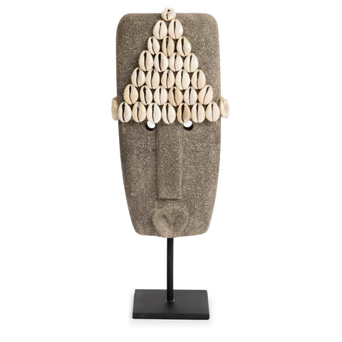 A hand-carved tribal mask sculpture with a stylized human face, adorned with a triangular arrangement of cowrie shells on top. Crafted from Sumba Stone, it stands elegantly mounted on a black stand.