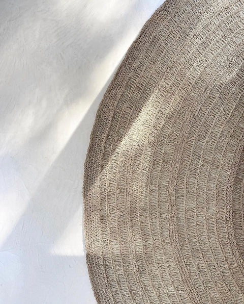 A round, woven jute rug lies on a light-colored floor, exuding the durability of a hardwearing seagrass carpet. Sunlight casts soft shadows across its surface, creating a warm and inviting atmosphere. The rug features a spiral pattern with textured details.