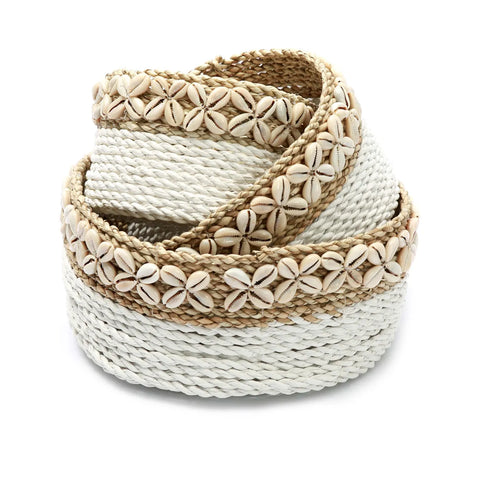 A decorative woven basket made of natural fibers, featuring a coiled design and an Ibiza vibe. It is embellished with rows of small, white cowrie shells, adding a textured and ornate appearance. This bohemian decorative item boasts a beige and white color scheme.