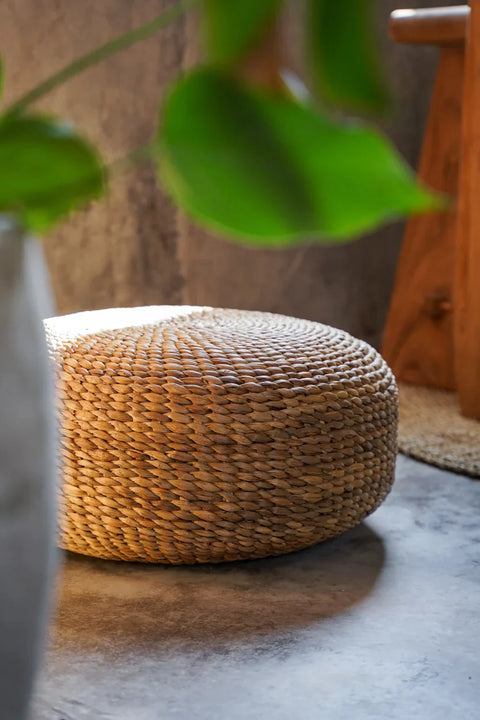 A round, water hyacinth pouf sits on a concrete floor, with a green plant in the foreground and a wooden stool in the background, enhancing the boho decor's cozy, natural ambiance.