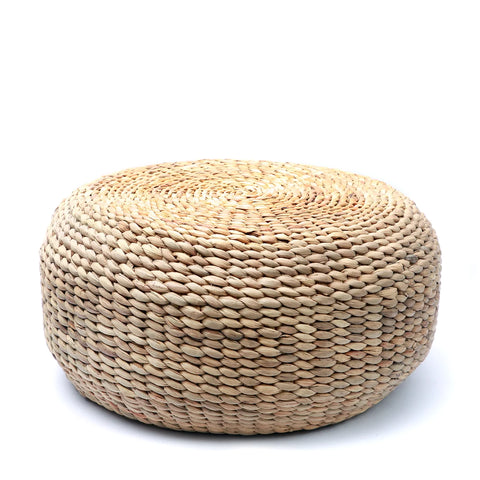 A round, boho-inspired pouf crafted from natural woven water hyacinth fibers features a textured surface, perfectly showcased against a plain white background.