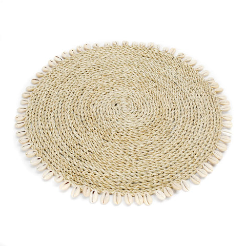 A round woven placemat with a spiral pattern made from natural fibers, this seagrass placemat is edged with cowrie seashells evenly spaced around the circumference, adding a touch of island style.