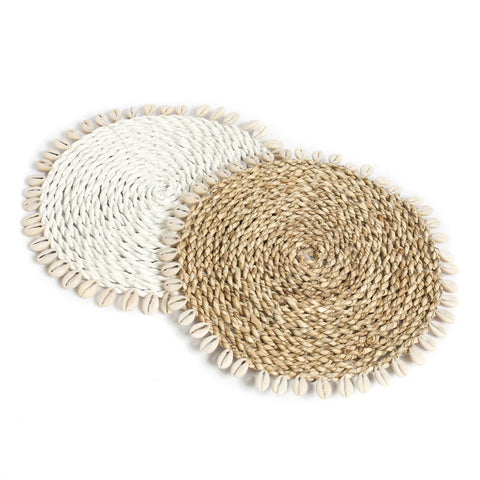 Two round woven placemats, one white and one natural, adorned with cowrie seashell borders, placed overlapping on a plain background. Perfect for adding a touch of island style to your table setting.