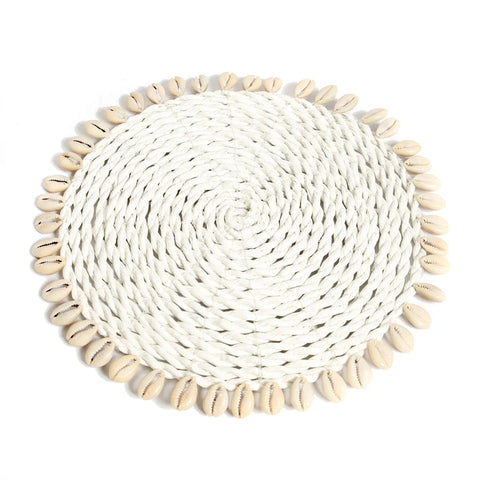 A circular white woven placemat adorned with cowrie seashells around the edge, arranged in a symmetrical pattern. The texture and natural color of the shells contrast beautifully with the simple woven material, evoking an island style charm.