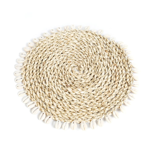 A round, woven wicker placemat with a spiral pattern, adorned with a border of small cowrie seashells, exudes an island style charm. The placemat rests gracefully on a plain white background.