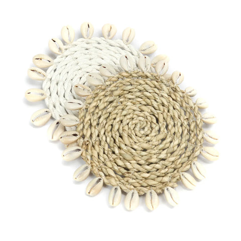 Two woven seagrass coasters, crafted from beige and white natural fibers, are adorned with small cowrie seashells around the edges. Stacked slightly overlapping each other on a white background, these coasters evoke an elegant island style.