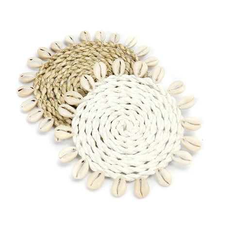 Two round, woven seagrass coasters made of natural fibers. One is light brown and the other is white, both capturing an island style essence. Each is beautifully embellished with a border of cowrie seashells around the edge.