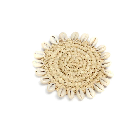 A round, woven Balinese coaster crafted from natural fibers features a circular pattern adorned with small white cowrie seashells around the edge, set against a white background.