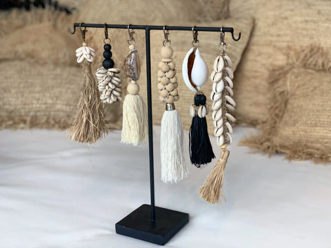 A display stand with eight boho luxury keychains featuring tassels, wooden beads, seashells, and macrame elements. The background showcases textured cushions in neutral tones.