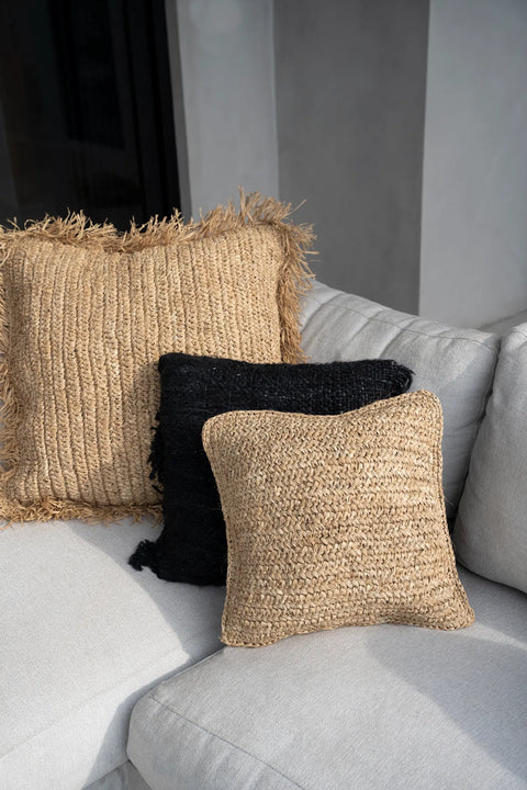 Three cushions are arranged on a light gray sofa. Two Raffia cushions, woven with natural, textured materials, radiate a boho style; the large one boasts fringe edges. A black cushion is tucked between them, adding contrast and enhancing the natural vibe of the arrangement.