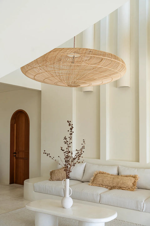 A modern living room featuring a large, handwoven hanging light fixture, a white sofa with textured raffia cushions, and a minimalist vase with branches on a sleek white table. The walls are light-colored with vertical accents, and a wooden door is in the background.