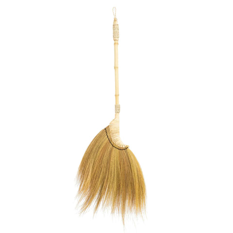 A traditional Indonesian broom with a wooden handle and straw bristles is gracefully suspended against a plain background. The bristles fan out from a woven section, and an attached loop makes it easy to hang as a decorative item.