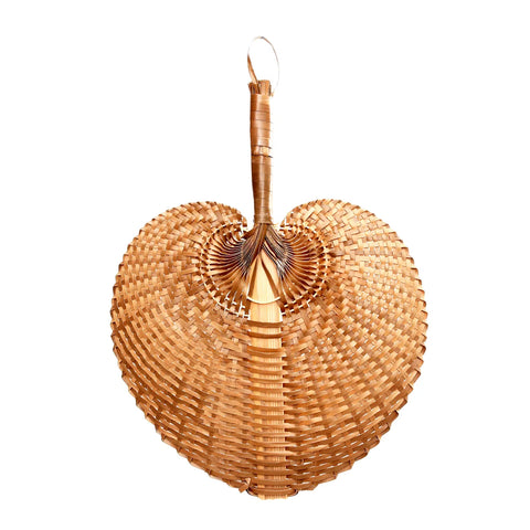 A handmade Balinese hand fan, heart-shaped and woven from natural straw, features intricate patterns crafted by local artisans. The fan, crafted with traditional techniques, includes a looped handle at the top for easy holding or hanging.