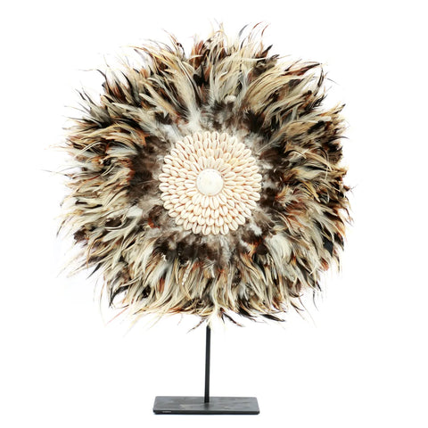 A bohemian beauty, this piece features a circle of brown, beige, and white feathers framing a central pattern of small shells. Elegantly mounted on a decorative stand, it's an exquisite brown feather display that captures the essence of natural artistry.