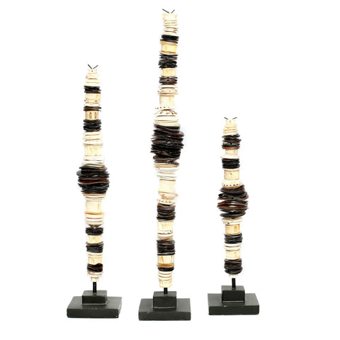 An eye-catching trio of abstract sculptures stands on black bases, evoking a coastal vibe. Each piece features vertically stacked circular elements in shades of brown, beige, and black, arranged in tall, slender forms. The varying heights add a touch of tribal luxe to the captivating display.