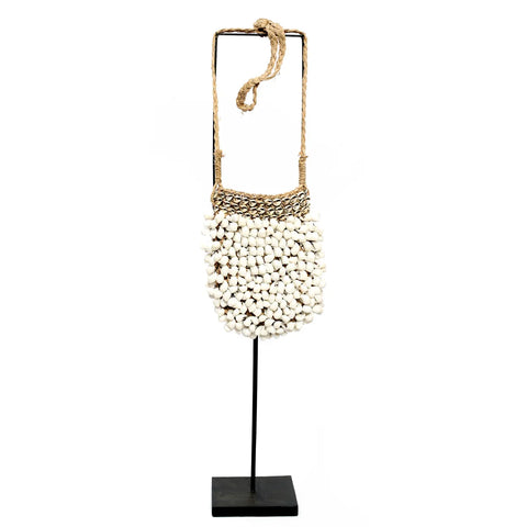 A white shell purse with a woven brown string strap is displayed on a black stand. This handmade bag is intricately designed with numerous shells, creating a textured appearance. The stand features a simple black square base, adding to its bohemian chic allure.