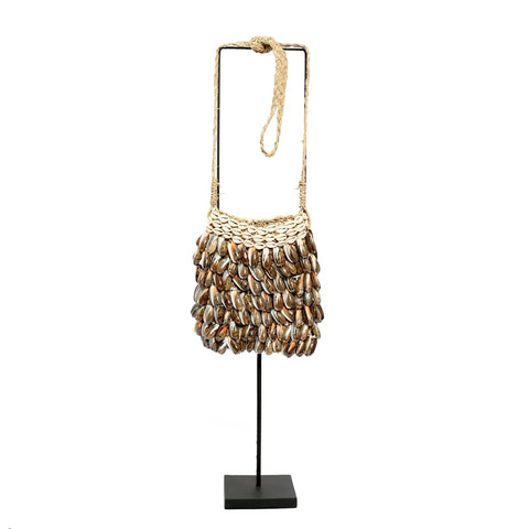 A bohemian chic shell purse made of woven material adorned with numerous seashells hangs from a stand. The intricately attached shells cover the entire handmade bag, and the strap appears to be crafted from braided fibers.