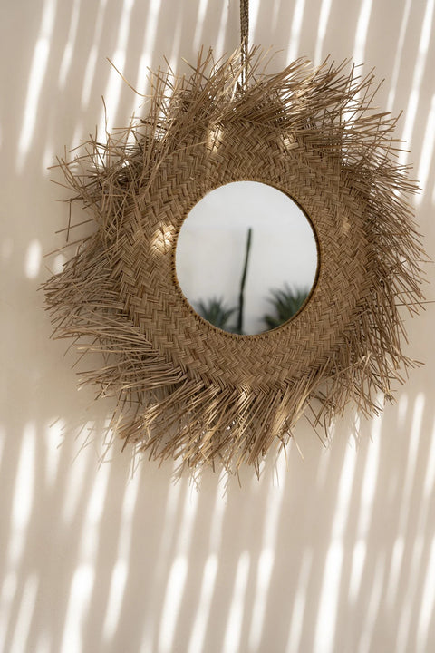 A round rattan wall mirror with a woven straw frame shaped like a sunburst hangs on the wall, adding a boho touch to the room. The mirror reflects faint images of indoor plants while sunlight creates striped shadows on the natural-colored walls' surface.