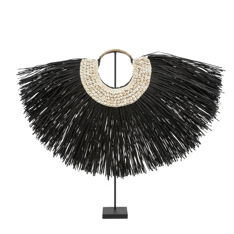 A bohemian beauty, this decorative piece showcases a large, circular fan-like shape with black fibers radiating outward. It's adorned with a crescent of white shells at the center and displayed elegantly on a metal stand, making it a striking alang grass display for any home interior.