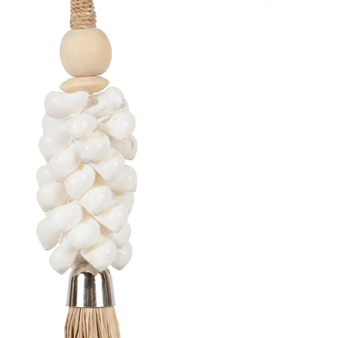 A decorative object with clustered cowrie shells topped with a wooden bead hangs from a braided rope. The bottom features a metallic cap with bristles, resembling a tassel. Crafted by Balinese artisans, it exudes a bohemian atmosphere against the plain white background.